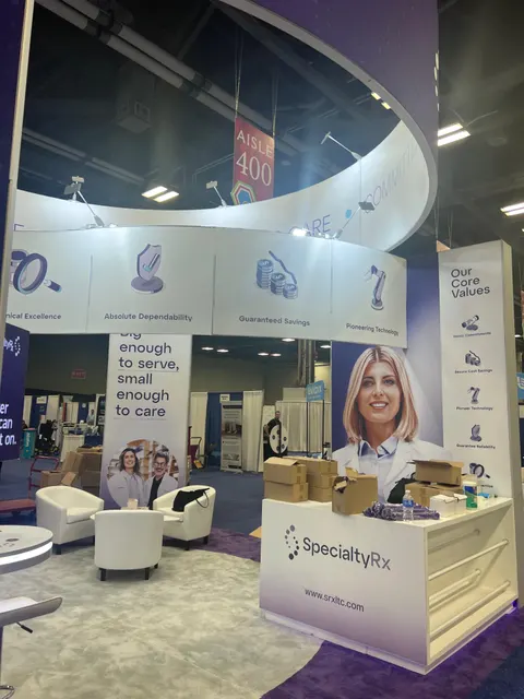 SpecialtyRx trade show booth built by Purple Exhibits: SpecialtyRx island booth with a large white curved overhead ring, purple accent carpet, a white reception counter with unpacked boxes on it and white lounge chairs,