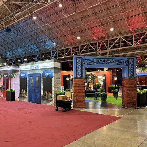 Amazon trade show booth built by Purple Exhibits: Blue 'Welcome to Amazon Quarter' archway on brick pillars leading into a courtyard with an olive tree, lounge seating and turf, beside a white brick facade with blue doub