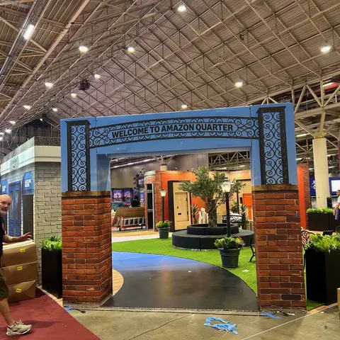 Amazon trade show booth built by Purple Exhibits: Close view of the blue and brick 'Welcome to Amazon Quarter' entrance arch with a curved black pathway, turf, olive tree and planters, with a worker and boxes visible at