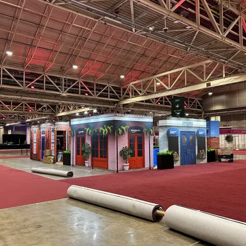 Amazon trade show booth built by Purple Exhibits: Wide view of the Amazon Quarter street-scene facades in brick with orange and blue doors and hanging plants, with rolls of red carpet still on the floor in the foreground