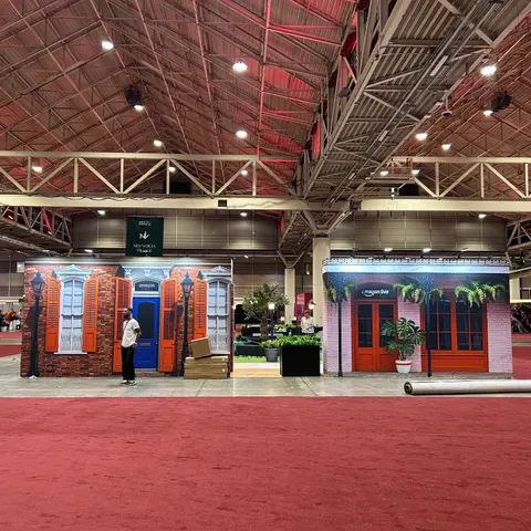 Amazon trade show booth built by Purple Exhibits: Front view of two Amazon Quarter storefront facades, one brick with orange shutters and one pink with green ferns and a red door, on red carpet with a worker and boxes at