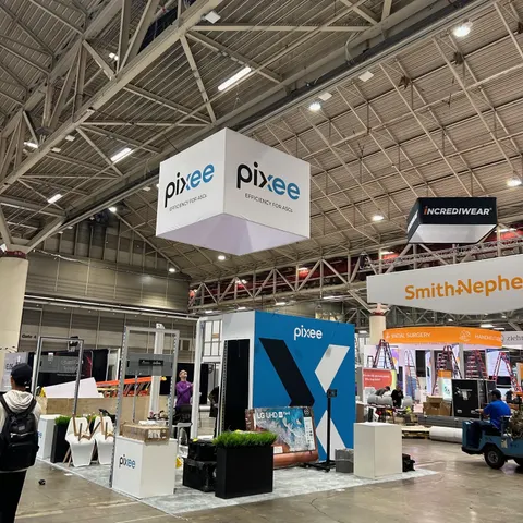 Pixee trade show booth built by Purple Exhibits: Pixee island booth still being set up on the show floor, with a large white square hanging sign, a bold blue backlit X-graphic wall, white counters and green planter boxes