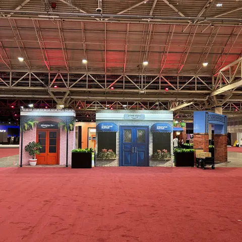 Amazon trade show booth built by Purple Exhibits: Wide, empty-hall view of an Amazon walk-through environment built as a row of themed storefronts with red and blue doors, brick facades, flower boxes and lanterns, standi