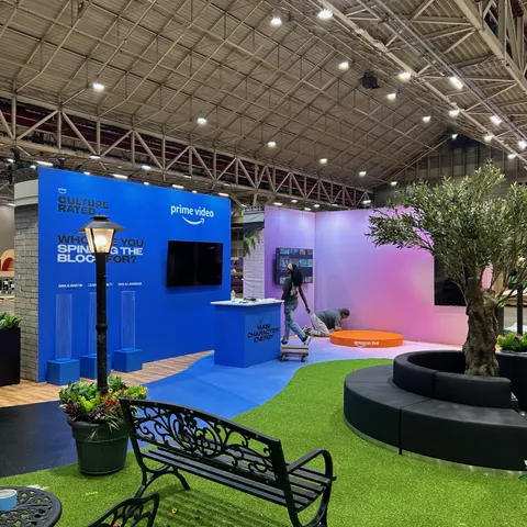 Prime Video trade show booth built by Purple Exhibits: Prime Video 'Culture Rated' activation with a bright blue graphic wall and screens, a pink-to-purple gradient curved wall, artificial olive tree with a round black l