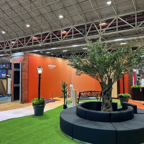 Amazon trade show booth built by Purple Exhibits: Amazon environment with a bold orange back wall bearing the Amazon logo, a red brick doorway, a large artificial olive tree in a round black lounge seat, green turf, blac