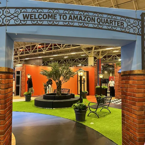 Amazon trade show booth built by Purple Exhibits: Entrance archway reading 'Welcome to Amazon Quarter' in wrought-iron style lettering over a blue frame with brick pillars, framing a view of the orange-walled town square