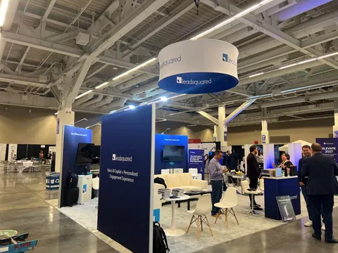 LeadSquared trade show booth built by Purple Exhibits: Navy and white LeadSquared island booth with a round hanging sign, screens on the walls, white lounge seating, white chairs and small meeting tables on a light grey