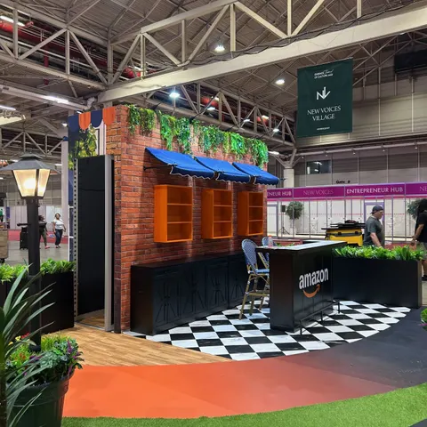 Amazon trade show booth built by Purple Exhibits: Amazon storefront-style environment with a red brick facade, orange shelving units under blue awnings, a black Amazon counter, bistro chair, street lamp, planters and bla
