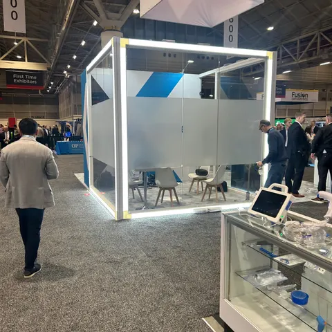 Trade show booth built by Purple Exhibits: Enclosed frosted-glass meeting cube with LED-edged frame and blue and white graphics, white chairs inside, a glass product case with medical devices in the foreground, visitors