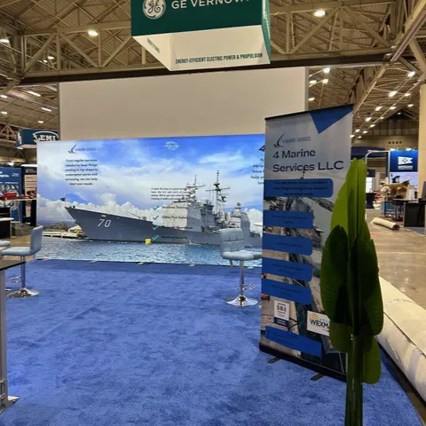 4 Marine Services LLC trade show booth built by Purple Exhibits: Inline booth for 4 Marine Services with a large backlit fabric wall showing a naval warship, a blue roll-up banner, white bar stools, blue carpet and an ar