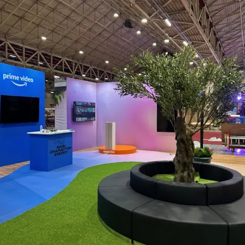 Prime Video trade show booth built by Purple Exhibits: Open Prime Video experiential space with a pink-to-blue gradient back wall, blue carpet, artificial turf, a large olive tree ringed by a circular black seating bench