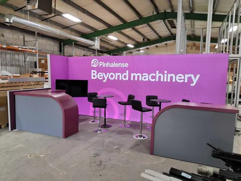 Pinhalense trade show booth built by Purple Exhibits: Pre-show warehouse setup of a magenta Pinhalense 'Beyond machinery' back wall with two curved grey-and-plum counters, black bar stools, high tables and a mounted scre