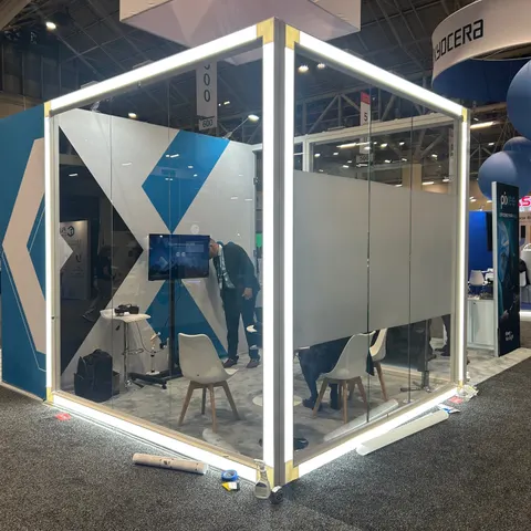 Pixee trade show booth built by Purple Exhibits: Close view of the Pixee LED-framed glass meeting cube with frosted panels, white chairs, a wall screen and a staffer inside; some setup debris on the grey carpet.