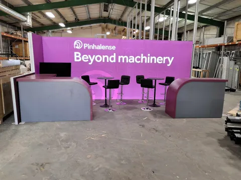 Pinhalense trade show booth built by Purple Exhibits: Straight-on warehouse preview of the magenta Pinhalense 'Beyond machinery' inline back wall with two curved plum counters, black stools, cocktail tables and a screen.