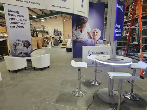 SpecialtyRx trade show booth built by Purple Exhibits: Warehouse staging of a SpecialtyRx pharmacy services booth with purple photo fabric graphics, a round illuminated white counter, white bar stools, cream armchairs an