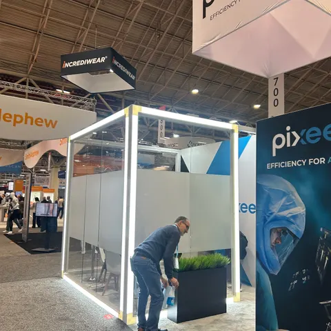 Pixee trade show booth built by Purple Exhibits: Side angle of the Pixee island showing the LED-framed glass meeting cube, a black planter with grass, a tall navy Pixee graphic tower and neighbouring Incrediwear hanging