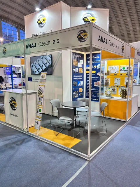 ANAJ Czech, a.s. trade show booth built by Purple Exhibits: Corner shell-scheme booth for ANAJ Czech with white and yellow panels, a wall screen, glass product showcase of gear hobs and cutting tools, grey chairs around