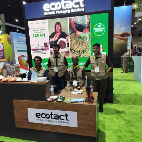 Ecotact trade show booth built by Purple Exhibits: Ecotact Hermetic Packaging Solutions inline booth with green photo graphics of coffee farming, a wood-grain reception counter, green turf flooring and four staff in khak