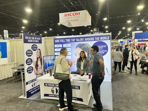 1 Source BPO trade show booth built by Purple Exhibits: Wider view of the 1 Source BPO 10x10 inline booth with blue and white talent-recruiting graphics, a white counter with printed literature, a monitor and staff talki
