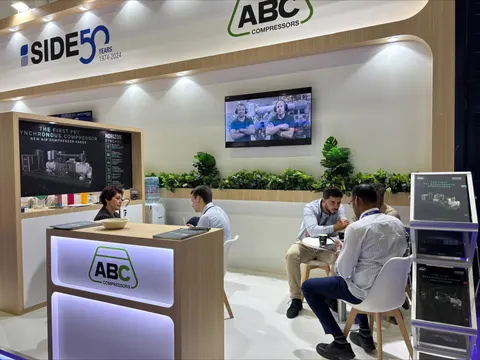 ABC Compressors trade show booth built by Purple Exhibits: Warm ABC Compressors booth celebrating 50 years of Side, with light-wood walls and counter, backlit logo, a wall TV, planted greenery shelves and white chairs wh