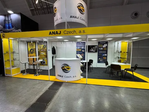 ANAJ Czech, a.s. trade show booth built by Purple Exhibits: Yellow-and-white inline booth for ANAJ Czech with a round hanging sign, glass display cases of metal parts and tools, a curved white reception counter and yello
