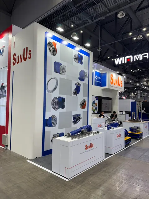 SunUs trade show booth built by Purple Exhibits: Tall white tower with a blue-and-grey graphic wall of gearboxes and hydraulic cylinders for SunUs, with blue mechanical components displayed on white plinths and staff at