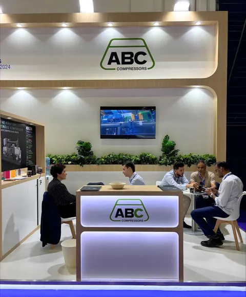 ABC Compressors trade show booth built by Purple Exhibits: Warm light-wood booth for ABC Compressors with a glowing backlit reception counter, a video screen, planter greenery and staff meeting visitors at a table.