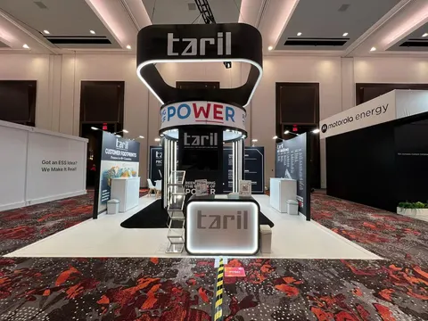 taril trade show booth built by Purple Exhibits: Black-and-white island booth for taril with an illuminated round hanging sign, a central tower with POWER graphics, backlit reception counter and screens on red patterned