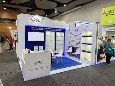 IMS trade show booth built by Purple Exhibits: Clean white and blue inline booth for IMS cast and machined metal components, with backlit shelving displaying small metal parts, a wall-mounted screen, branded counter, whi