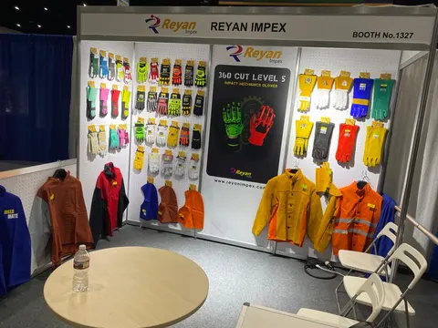 Reyan Impex trade show booth built by Purple Exhibits: Inline booth 1327 for Reyan Impex safety gloves and workwear, with colourful gloves pegged on white walls, hanging hi-vis jackets, a round wooden table and grey carp