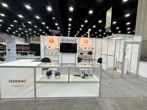 Sedemac trade show booth built by Purple Exhibits: White inline booth for Sedemac innovative controls with glass display cases of engine control units, a TV screen, shelves and an enclosed meeting room with grey carpet.