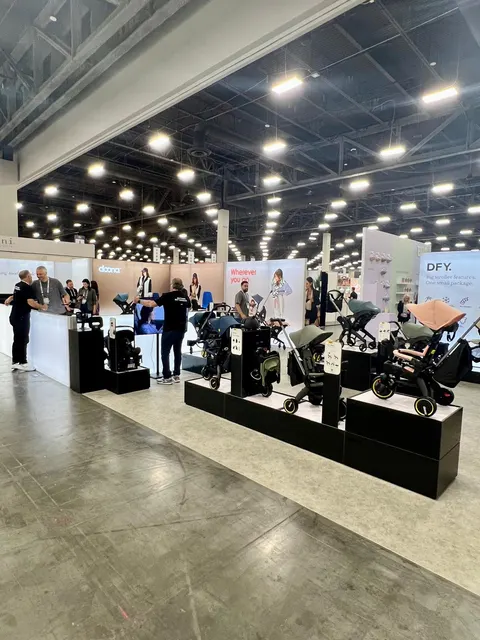 Doona trade show booth built by Purple Exhibits: Open Doona island booth with strollers and car seats displayed on black plinths, soft pink and beige lifestyle graphics on the back wall and staff talking with visitors.