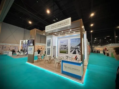 Eagle Chemicals trade show booth built by Purple Exhibits: Eagle Chemicals island booth with light wood walls, backlit header, factory photo panels, white bistro seating, a blue reception counter and glowing teal carpet.