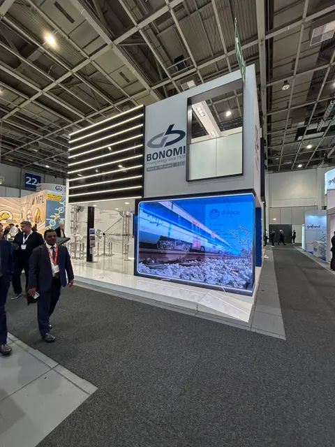 Bonomi trade show booth built by Purple Exhibits: Two-storey white Bonomi booth with a huge LED wall showing a train image, slatted lit ceiling detail, upper-level meeting space and visitors on the grey aisle carpet.