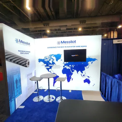 Messilot trade show booth built by Purple Exhibits: Inline booth with backlit white walls, blue world map graphic, wall-mounted TV, three bar tables with stools and blue carpet for Messilot elevator wire ropes.