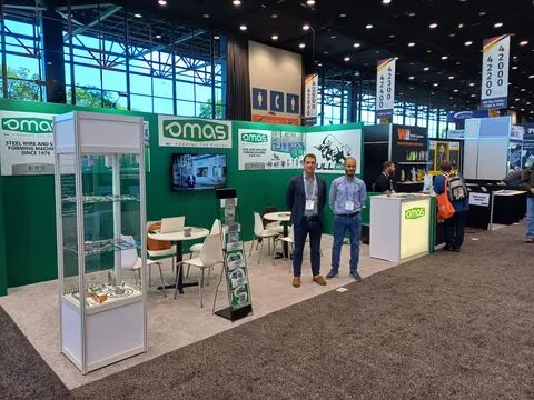 OMAS trade show booth built by Purple Exhibits: Green and white inline booth for OMAS steel wire forming machinery with a glass showcase, TV screen, bistro tables, a rounded counter and two staff standing.