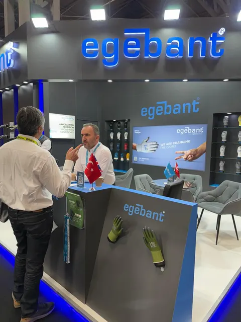 egebant trade show booth built by Purple Exhibits: Grey egebant booth with backlit logo, angled reception counter showing green gloves, LED screen and grey lounge chairs, with staff talking to a visitor.