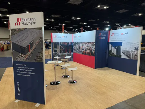 Ziemann Holvrieka trade show booth built by Purple Exhibits: Empty inline booth with a tall navy Ziemann Holvrieka tower panel, red and navy brewery-facility graphics on the back wall, white bistro tables with chrome sto