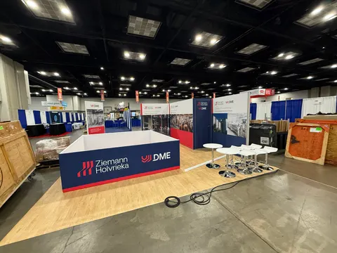 Ziemann Holvrieka / DME trade show booth built by Purple Exhibits: Wide pre-show view of the Ziemann Holvrieka and DME inline booth with a red and navy branded counter, back-wall graphics, white bistro tables on wood-loo