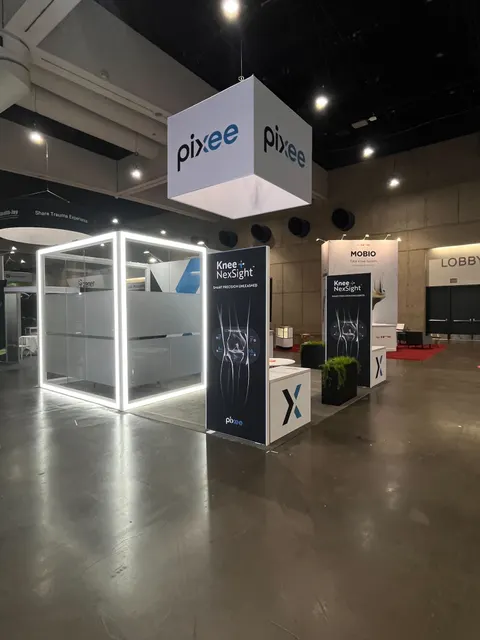 Pixee trade show booth built by Purple Exhibits: Pixee island booth with a white cube hanging sign, an LED-edge-lit glowing frame structure, navy Knee+ NexSight graphic towers, white counters with X logos and black plant