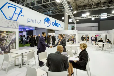 Olon / Infa Group trade show booth built by Purple Exhibits: White Olon 'part of Infa Group' booth with blue branding, white meeting tables and chairs, and industrial photo graphics, busy with visitors shaking hands and