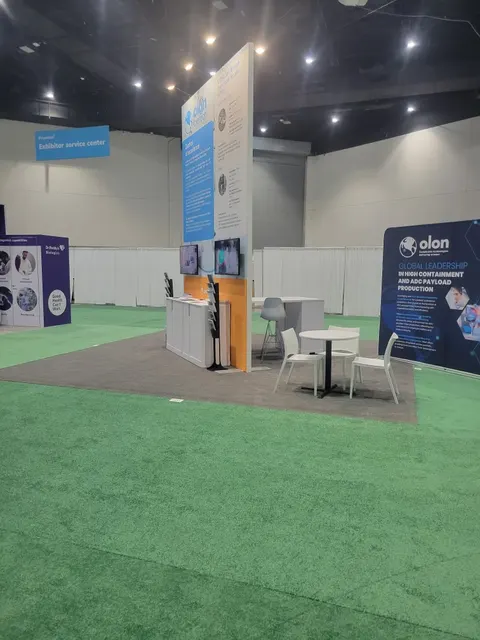 Olon trade show booth built by Purple Exhibits: Small Olon inline booth with a tall white tower with blue graphics, orange wood-accent counter with two screens, a white table and chairs, and a navy 'Global Leadership' ba