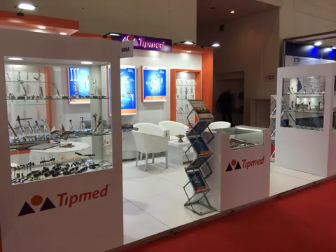 Tipmed trade show booth built by Purple Exhibits: Tipmed corner booth with orange and white walls, glass display cases of orthopaedic implants and instruments, backlit product posters, white armchairs and a branded count