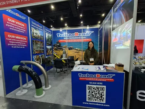 TechnoBend trade show booth built by Purple Exhibits: TechnoBend inline booth with blue and red pipeline-bend graphics, a green-and-grey pipe bend sample display, a branded counter with QR code, a screen, and a staff mem