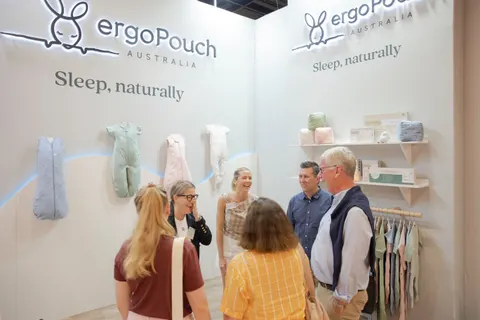 ergoPouch trade show booth built by Purple Exhibits: Visitors chatting inside the white ergoPouch booth with pastel sleep sacks on the wall, product shelving and a garment rail in the background.