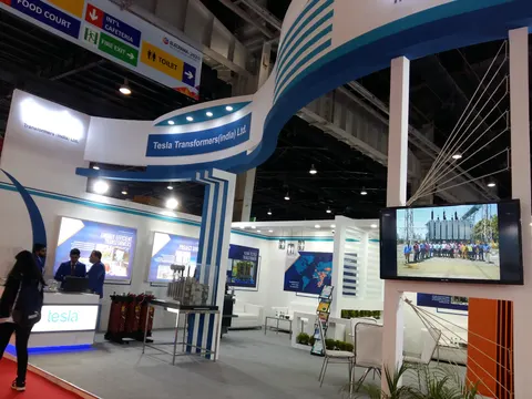 Tesla Transformers (India) Ltd. trade show booth built by Purple Exhibits: Wide view of the white and blue Tesla Transformers India island booth with a sweeping curved overhead ribbon, TV screens, transformer units on di