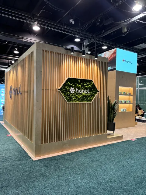 hanul trade show booth built by Purple Exhibits: Striking warm-wood slatted island booth for hanul with a hexagonal moss-wall logo feature, spotlights, backlit product shelving niche, potted plants, an overhead LED scree
