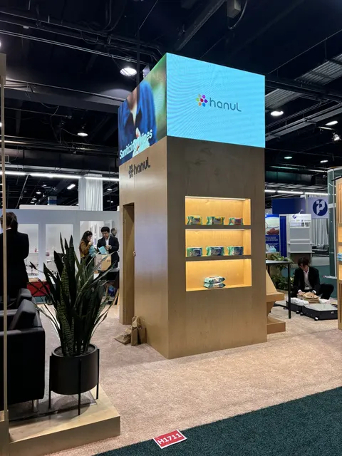 Hanul trade show booth built by Purple Exhibits: Tall plywood-clad tower with a wraparound LED header showing the Hanul logo, backlit product shelves displaying packaged goods, a black lounge chair and a large snake plan