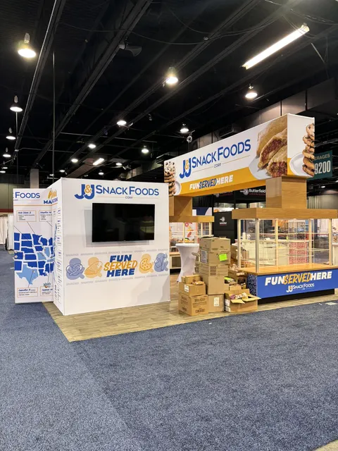 J&J Snack Foods trade show booth built by Purple Exhibits: White and gold J&J Snack Foods booth with a large TV wall, a pretzel-graphic header above a wood-clad sampling kiosk and a US map graphic panel; cardboard boxes 