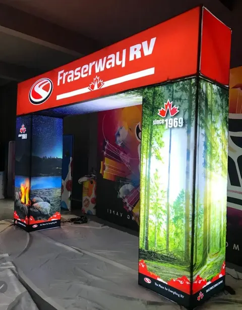 Fraserway RV trade show booth built by Purple Exhibits: Bright red Fraserway RV backlit archway with a forest-scene lightbox pillar and a lakeside campfire pillar, photographed during pre-show setup on a tarp-covered flo